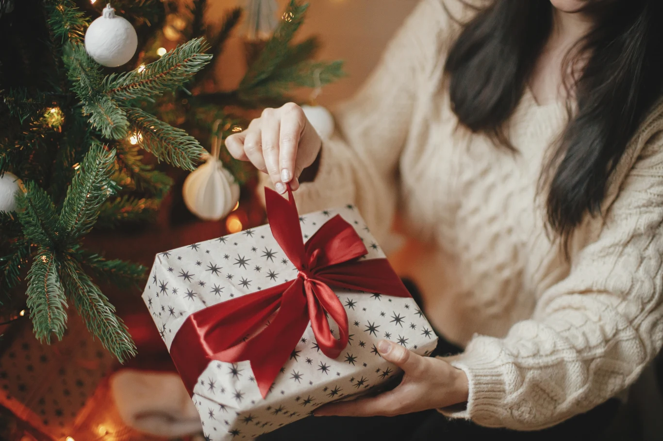 Kupując prezent, dobrze się zastanów, czy nie zranisz czyichś uczuć Osoba w jasnym swetrze trzyma elegancko zapakowany prezent z czerwoną wstążką, obok udekorowana choinka z jasnymi bombkami i świątecznymi światełkami.