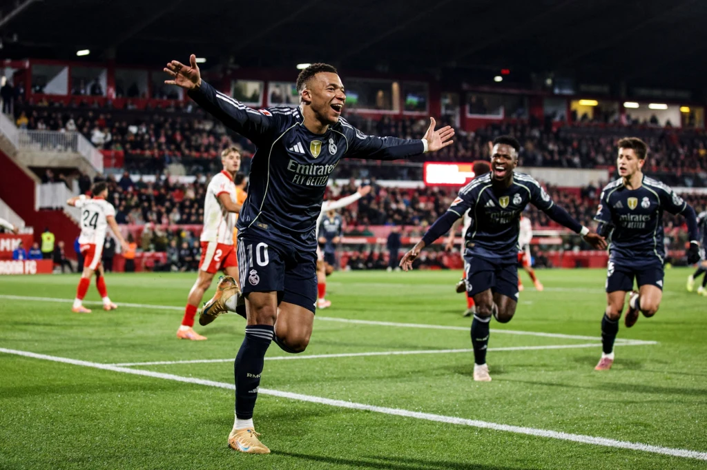 Kylian Mbappe, Vinicius Junior i Arda Guler to największe gwiazdy Realu Madryt w tym sezonie Piłkarz w ciemnym stroju rozpościera ręce w geście triumfu po zdobyciu gola podczas meczu na stadionie, a za nim biegną radośni koledzy z drużyny na tle pełnych trybun.