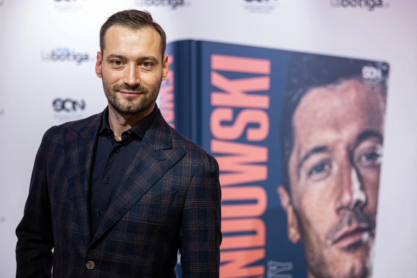 Mężczyzna w eleganckiej, ciemnej marynarce stoi przed dużym plakatem książki z widoczną okładką, na której znajduje się portret mężczyzny oraz pionowo napisane nazwisko.