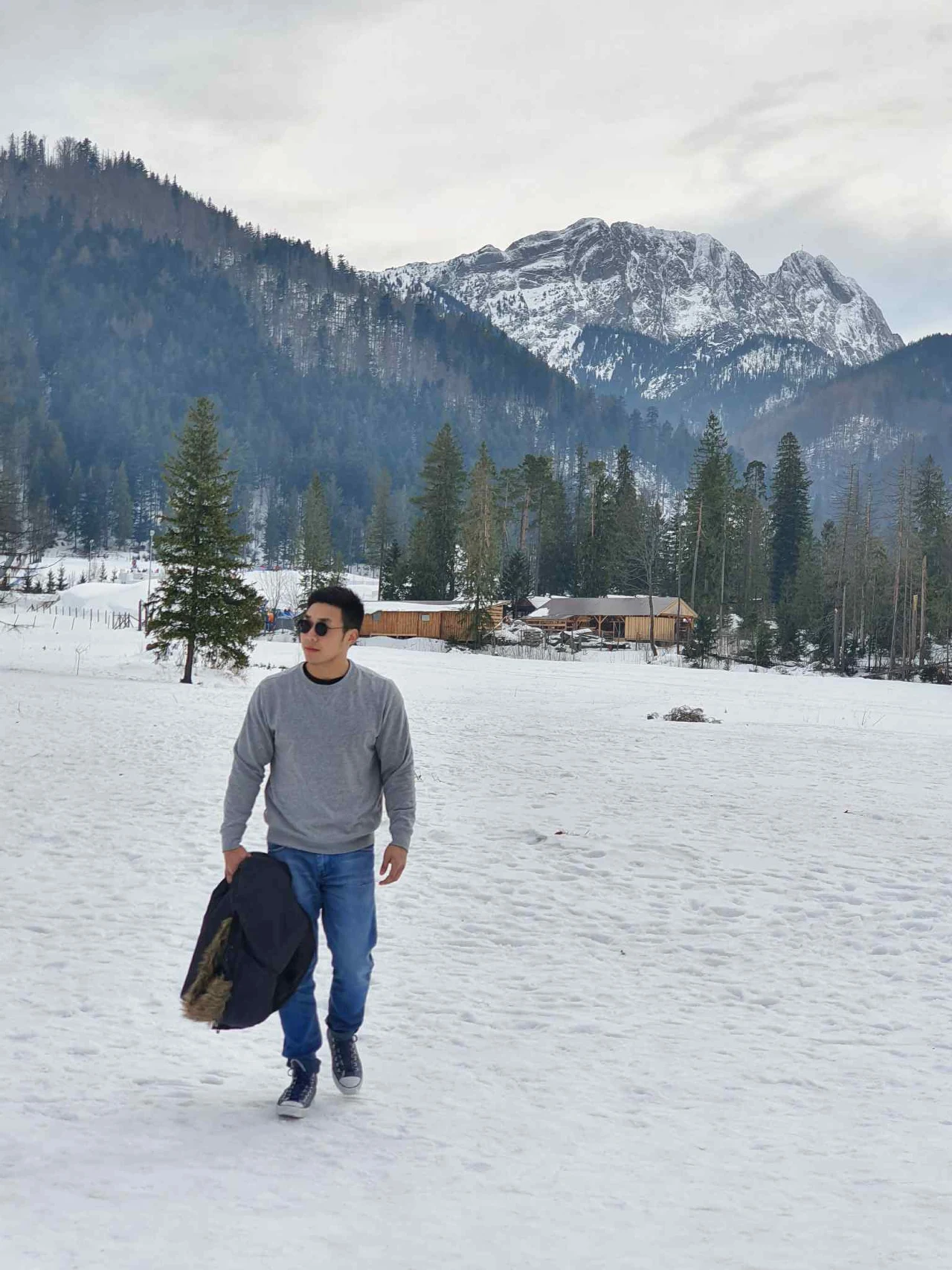 Mężczyzna spacerujący po śnieżnej polanie z kurtką w ręku, w tle ośnieżone góry, las oraz drewniane domki.