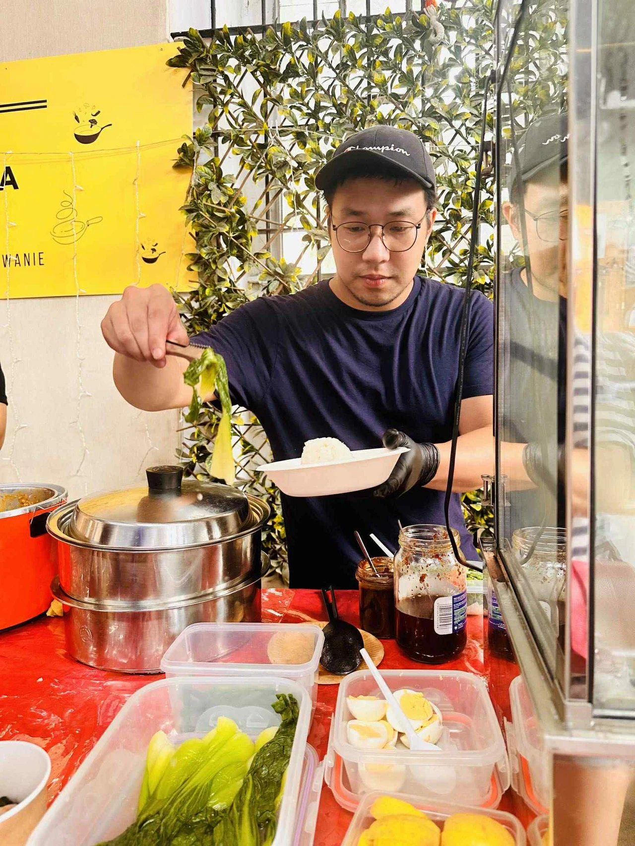 Mężczyzna w okularach i czapce, stojący za stołem z jedzeniem, przygotowuje posiłek, nakładając warzywa do plastikowego pojemnika. Na stole znajdują się ugotowane jajka, świeże warzywa, słoiki z sosami oraz metalowy garnek. W tle jasny żółty plakat i o...