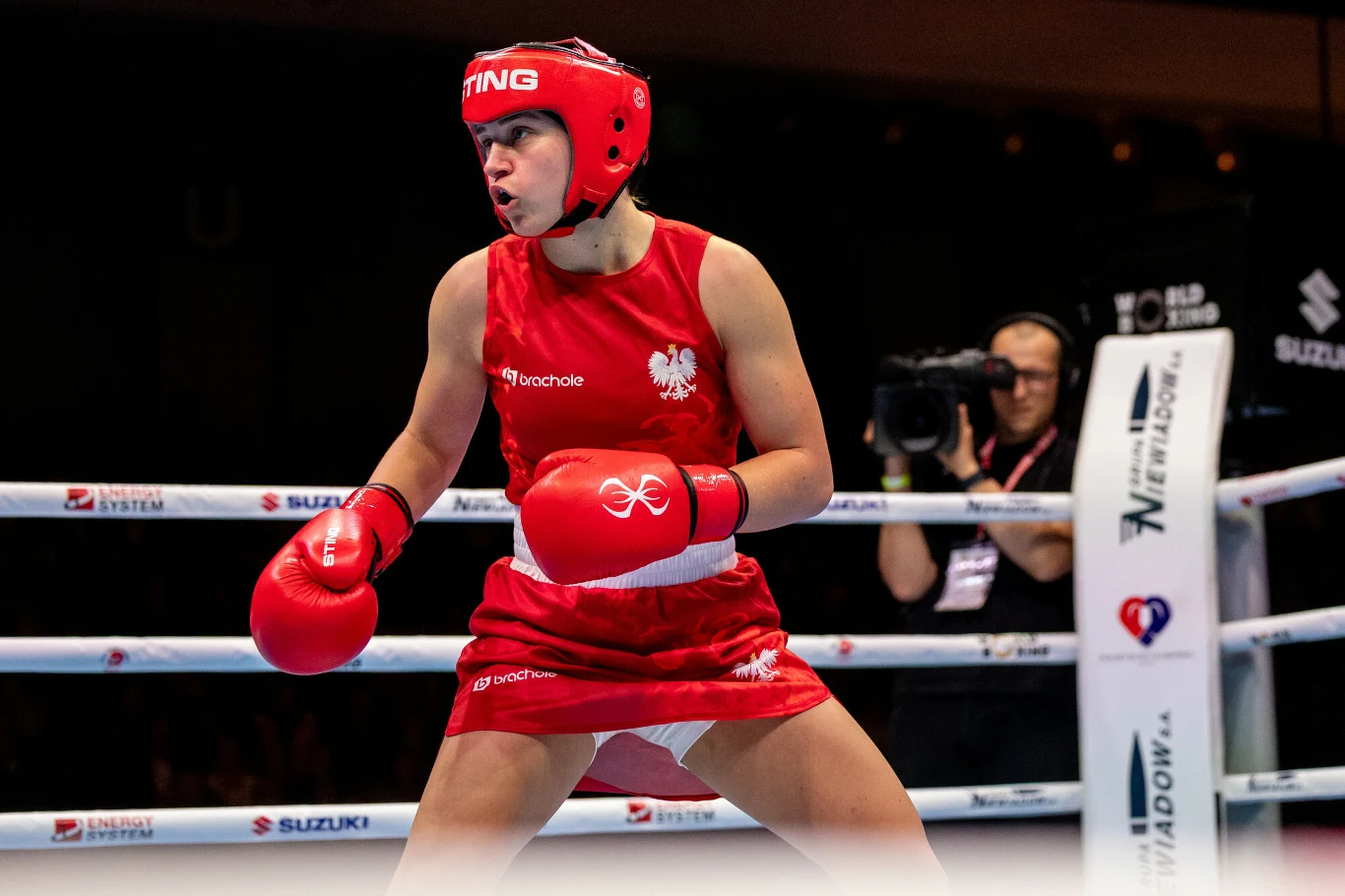 Julia Szeremeta Zawodniczka w czerwonym stroju bokserskim z herbem Polski na piersi przygotowuje się do walki na ringu, w tle widoczny jest operator kamery rejestrujący pojedynek.