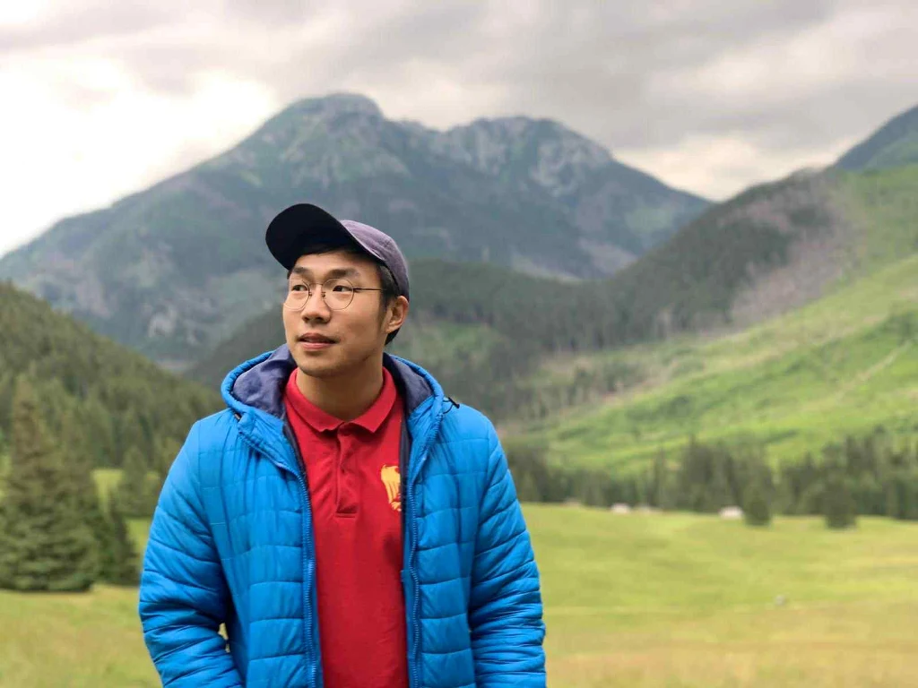 Mężczyzna w niebieskiej kurtce i czerwonej koszuli stoi na tle zielonych łąk i gór, w tle widoczne są drzewa oraz szczyty górskie pod pochmurnym niebem.