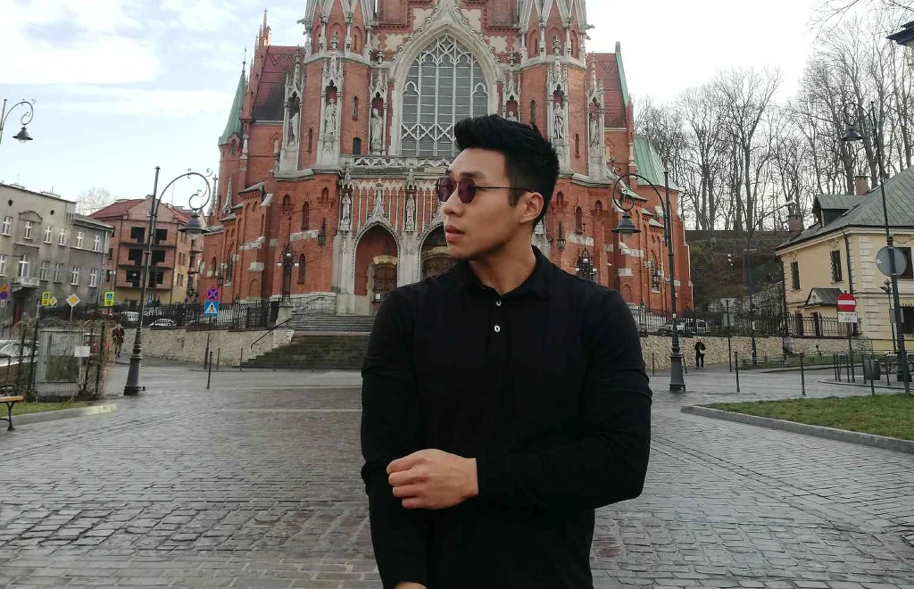 Mężczyzna w czarnej koszuli stoi na tle gotyckiego kościoła z czerwonej cegły. Spogląda w lewo, ma na sobie okulary, a wokół niego znajduje się pusty plac i zabudowa miejska.