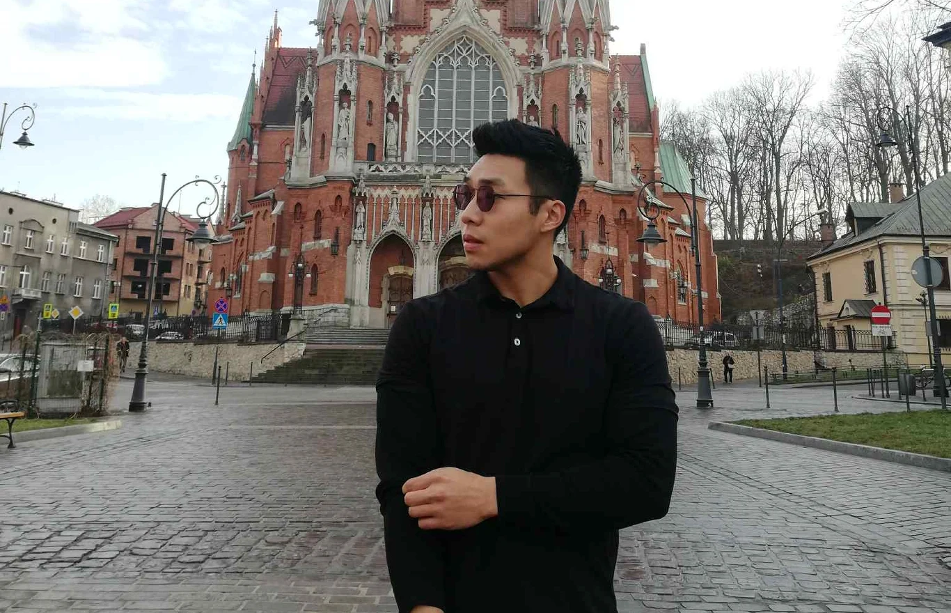 Mężczyzna w czarnej koszuli stoi na tle gotyckiego kościoła z czerwonej cegły. Spogląda w lewo, ma na sobie okulary, a wokół niego znajduje się pusty plac i zabudowa miejska.