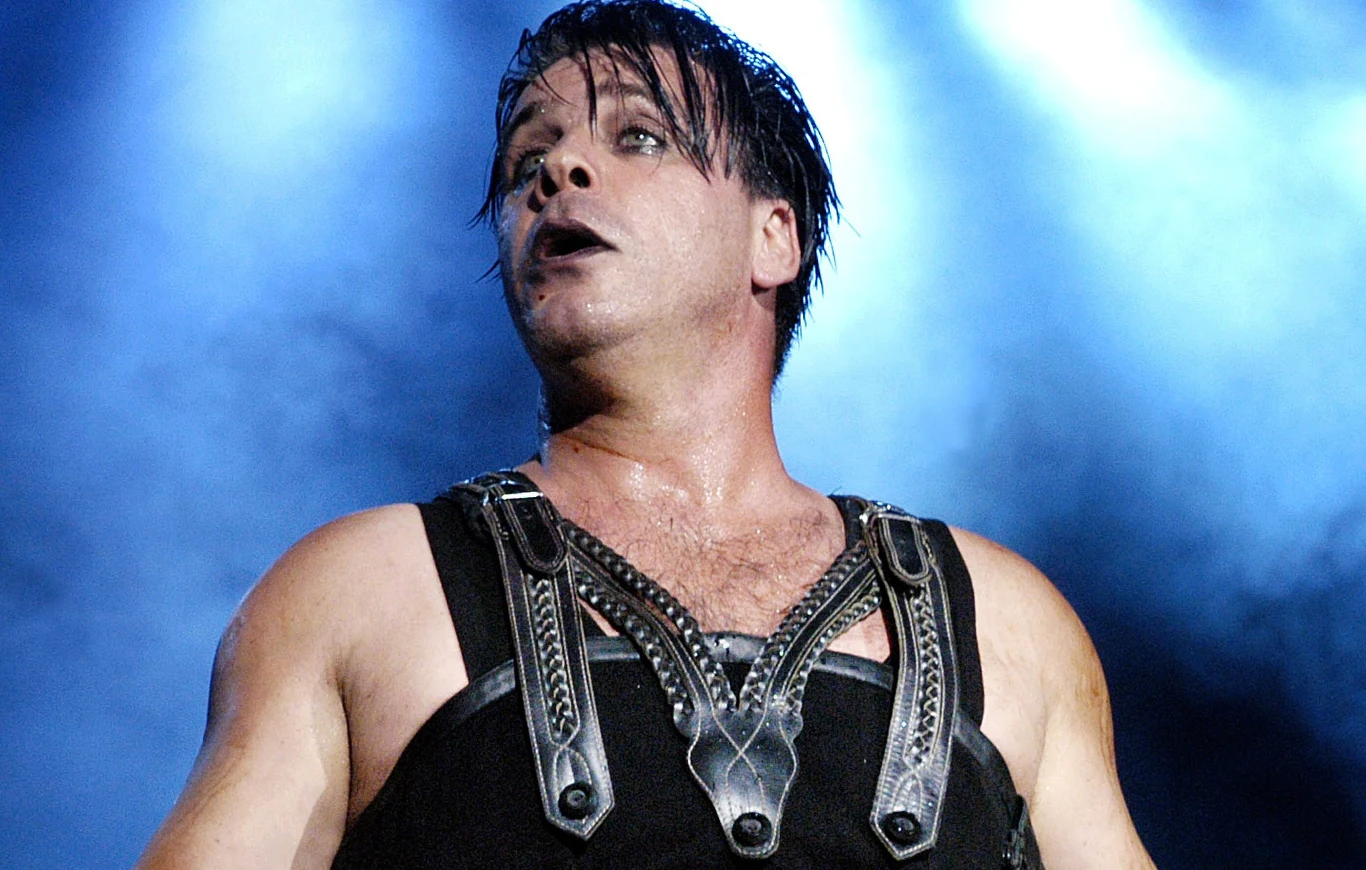 Till Lindemann wystąpił w Krakowie Mężczyzna w czarnej skórzanej kamizelce z metalowymi elementami, ciemny makijaż wokół oczu i ust, intensywne oświetlenie sceniczne w tle tworzące wyrazisty kontrast.
