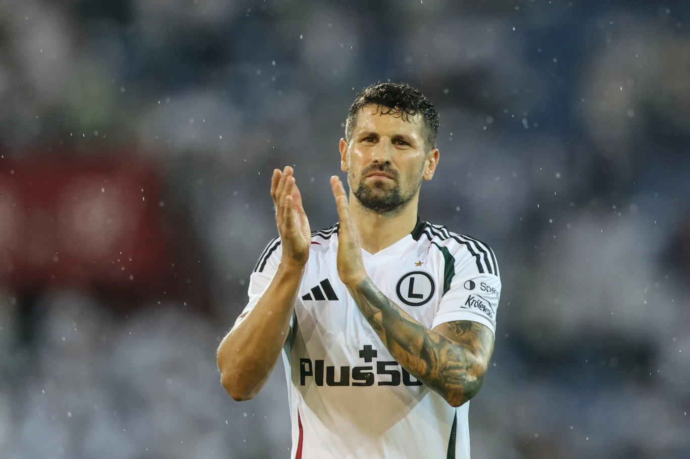 Paweł Wszołek Piłkarz w stroju meczowym Legii Warszawa oklaskuje, stojąc na tle rozmytego tłumu kibiców.