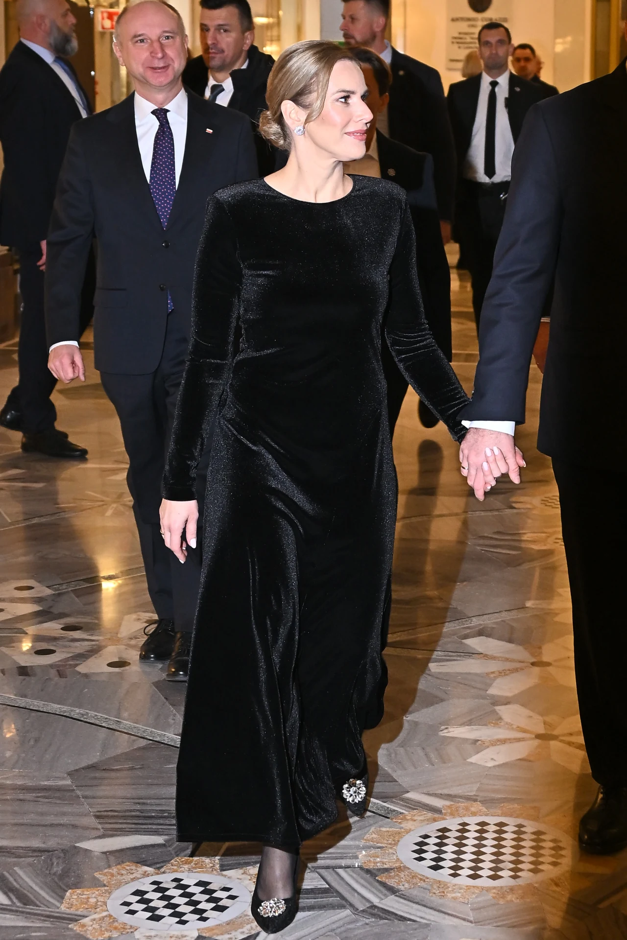 Elegancko ubrana kobieta w czarnej sukni idzie korytarzem w towarzystwie mężczyzn w garniturach, w otoczeniu ochrony i osób towarzyszących, w reprezentacyjnym wnętrzu o marmurowej podłodze.