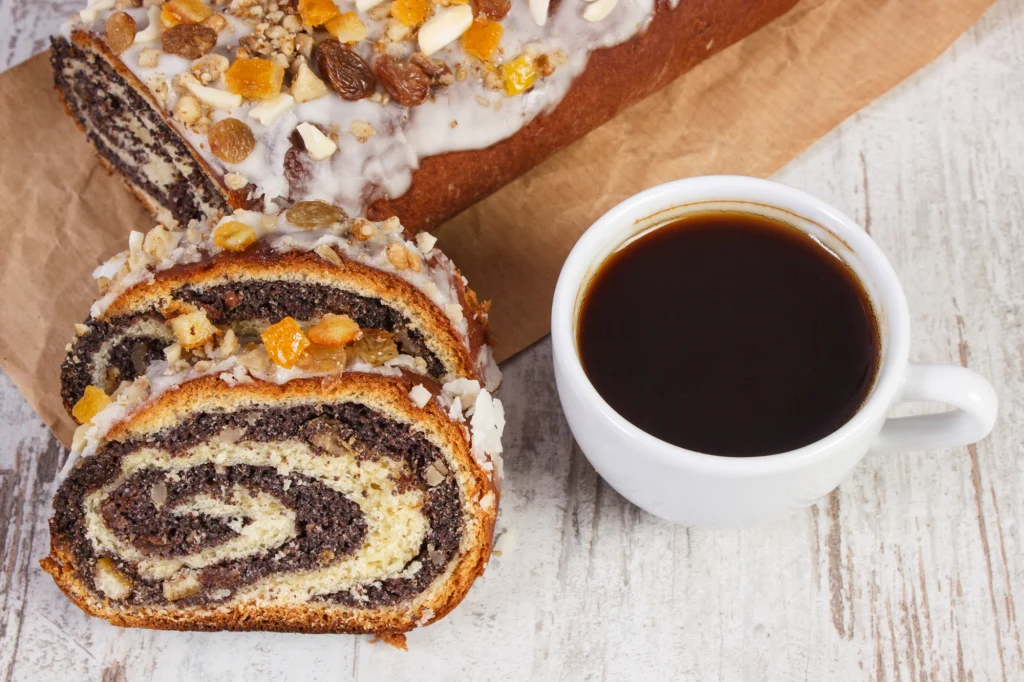 Kawa w białej filiżance obok świeżo pokrojonego makowca udekorowanego orzechami i suszonymi owocami, ustawionego na pergaminie na jasnym drewnianym stole.