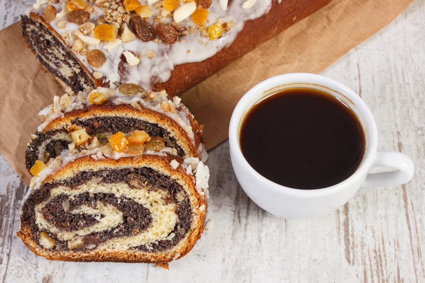 Kawa w białej filiżance obok świeżo pokrojonego makowca udekorowanego orzechami i suszonymi owocami, ustawionego na pergaminie na jasnym drewnianym stole.