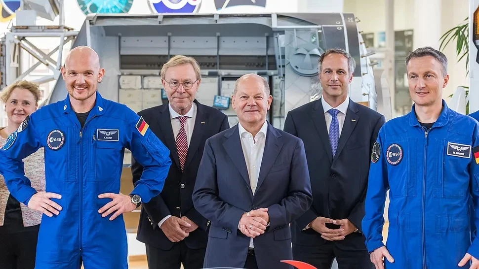 Grupa sześciu osób uśmiechających się do aparatu, w tym trzech mężczyzn w niebieskich kombinezonach astronautów ESA, dwóch mężczyzn w garniturach oraz kobieta. W tle fragment sprzętu kosmicznego na wystawie w nowoczesnym pomieszczeniu.
