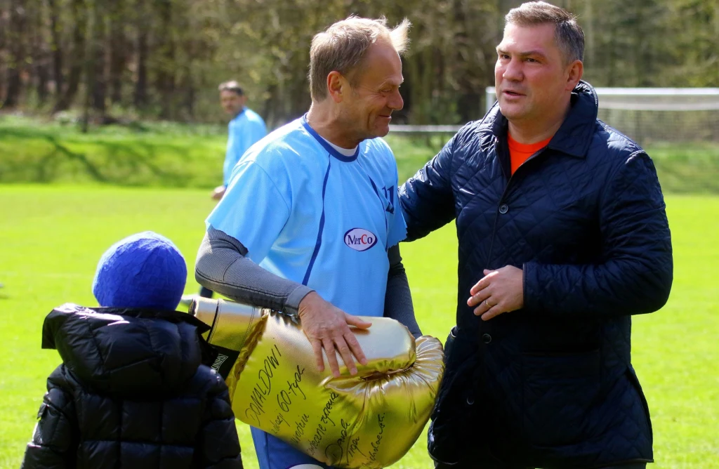 Dariusz Michalczewski wręczył Donaldowi Tuskowi złotą rękawice z okazji 60. urodzin premiera Dwaj mężczyźni rozmawiają na boisku piłkarskim, jeden z nich trzyma dużego, złotego balona w kształcie pucharu z widocznymi napisami, obok stoi dziecko w granatowej kurtce i niebieskiej czapce.