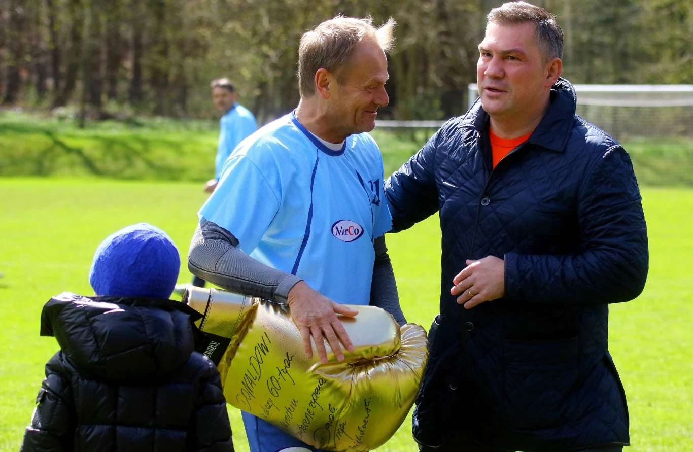 Dwaj mężczyźni rozmawiają na boisku piłkarskim, jeden z nich trzyma dużego, złotego balona w kształcie pucharu z widocznymi napisami, obok stoi dziecko w granatowej kurtce i niebieskiej czapce.