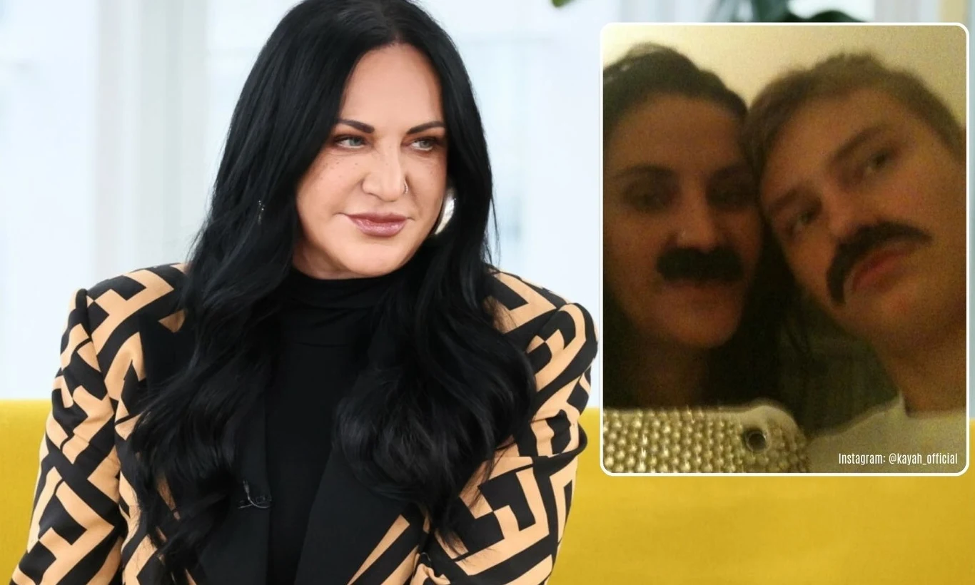 Kobieta z ciemnymi, długimi włosami siedzi na żółtej sofie ubrana w czarno-beżową marynarkę o geometrycznym wzorze, w tle jasne wnętrze. W prawym górnym rogu znajduje się wstawka z rozmytym zdjęciem dwóch osób, które mają doklejone lub narysowane wąsy.