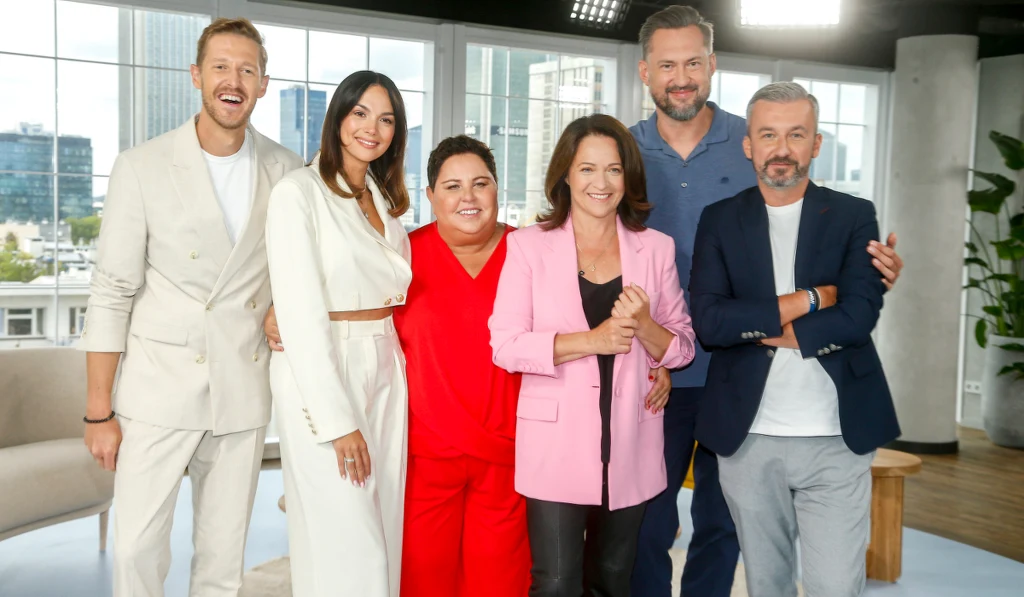 Grupa sześciu uśmiechniętych osób ubranych elegancko, pozujących razem w jasnym nowoczesnym wnętrzu z dużymi oknami i widokiem na miasto w tle.