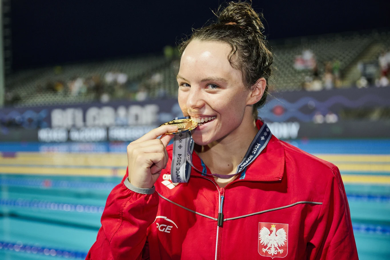 Sportowczyni w czerwonym dresie z godłem Polski na piersi pozuje na tle basenu, trzymając w ustach złoty medal zwisający na wstążce.