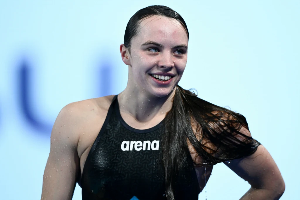 Młoda kobieta w stroju pływackim, z mokrymi włosami, uśmiecha się szeroko stojąc na tle rozmytego jasnego tła.