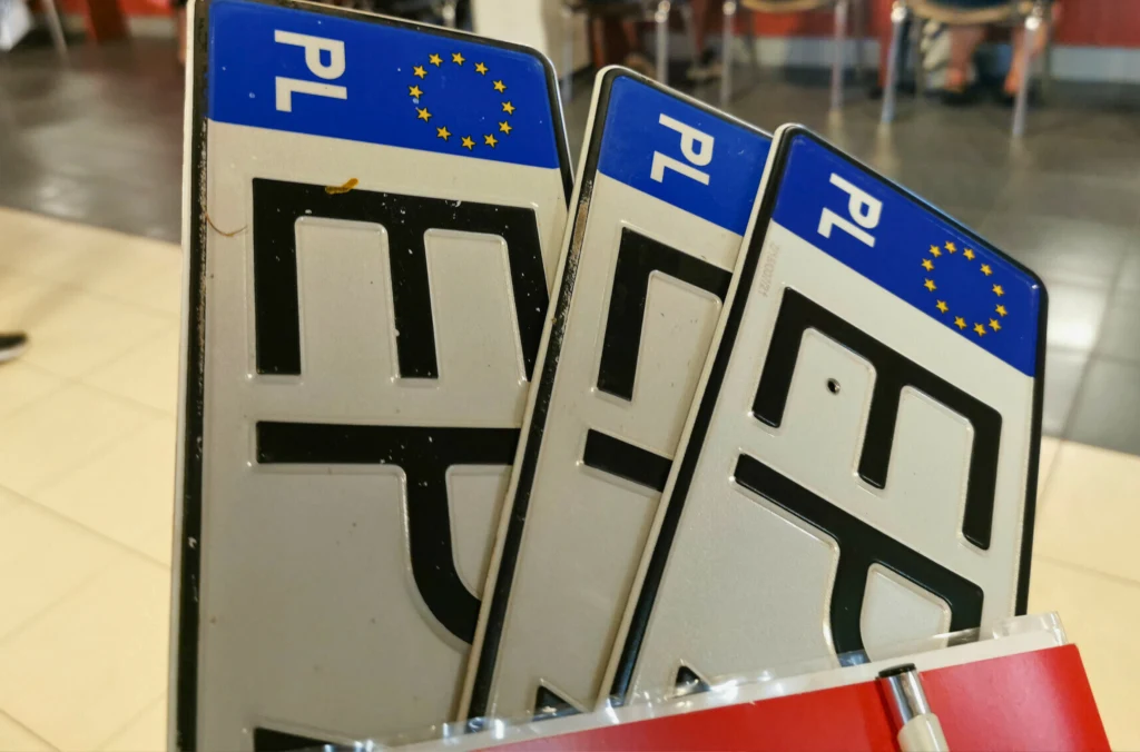 Trzy polskie tablice rejestracyjne z literami na białym tle z błękitnym paskiem po lewej stronie, zawierającym symbol Unii Europejskiej oraz oznaczenie kraju PL, ustawione ukośnie obok siebie na tle jasnej posadzki i czerwonego elementu.