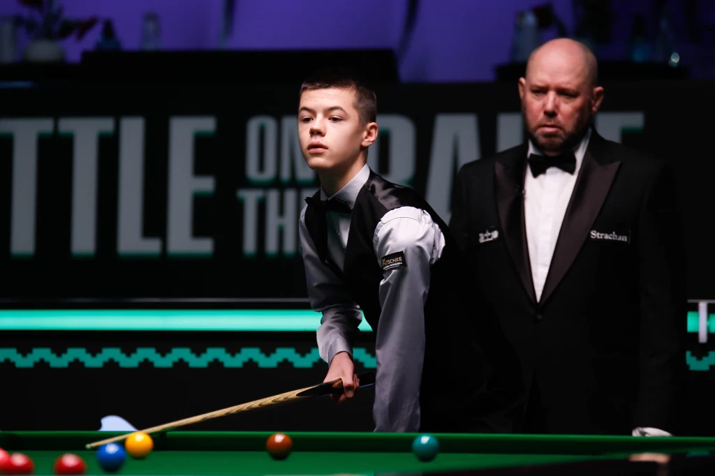 Młody snookerzysta w eleganckiej kamizelce wykonuje ruch przy stole bilardowym, obok stoi sędzia w czarnym garniturze. Tło ciemne, na ścianie widoczny duży napis promujący wydarzenie sportowe.