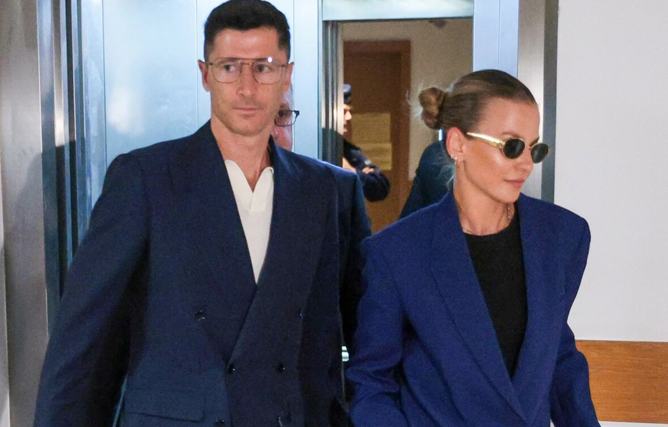 Para ubrana w eleganckie granatowe garnitury, wychodząca wspólnie przez drzwi windy. Kobieta ma związane włosy i okulary przeciwsłoneczne, mężczyzna nosi okulary i również garnitur. Tło to wnętrze budynku z neutralnym oświetleniem.