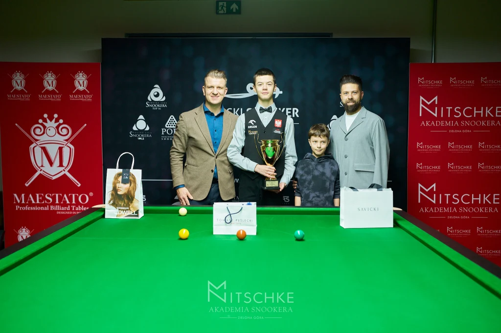 Grupa czterech osób pozuje przy stole snookerowym, jedna z nich trzyma puchar, wokół na stole ustawione nagrody i piłki bilardowe, w tle widoczne logotypy sponsorów i banery akademii snookera.