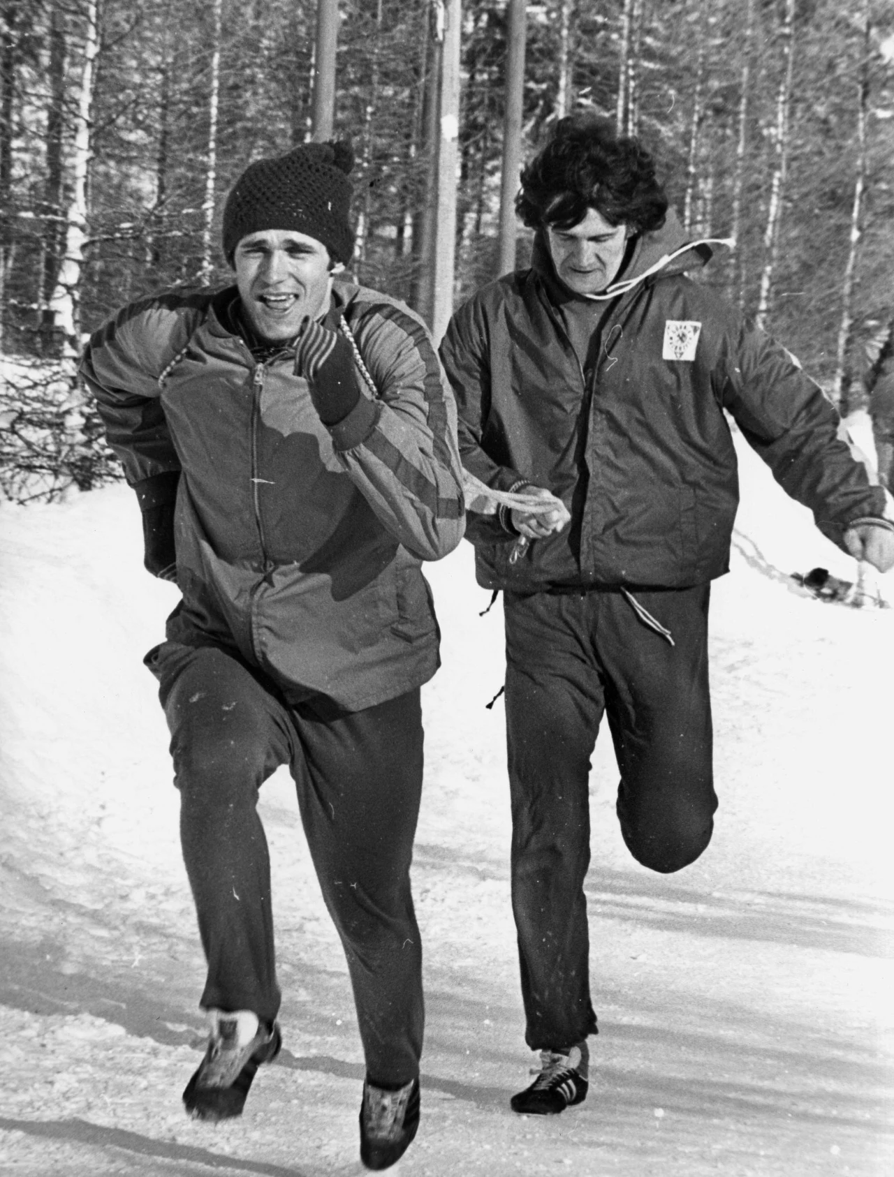 Dwóch mężczyzn w sportowych ubraniach biegnie ścieżką w zaśnieżonym lesie, pierwszy z nich skupiony jest na wysiłku, drugi podąża tuż za nim. Obaj ubrani są w kurtki i spodnie dresowe, w tle widoczny jest zimowy krajobraz z drzewami i śniegiem.