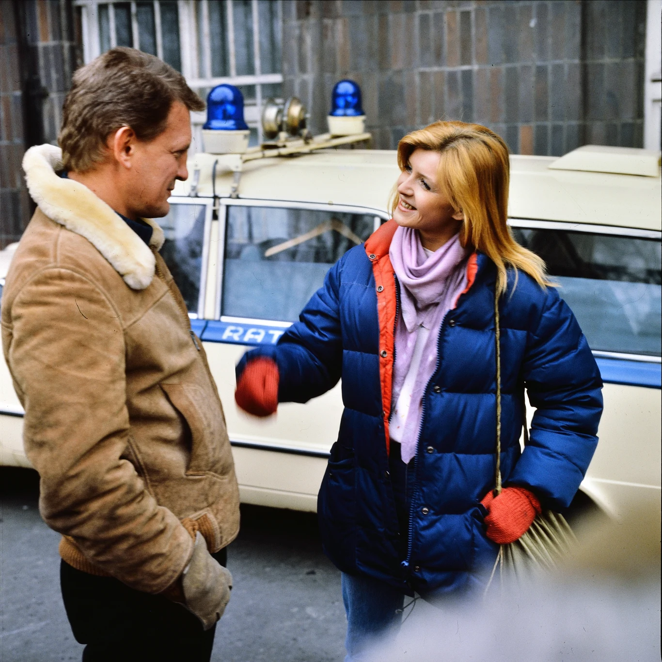 Maria Probosz na planie "07 zgłoś się" Kobieta w niebieskiej kurtce i czerwonych rękawiczkach rozmawia z uśmiechem z mężczyzną w brązowej kurtce, stojąc przed samochodem ratowniczym z dwoma niebieskimi sygnałami świetlnymi. W tle widać budynek oraz metalowe kraty na oknach.