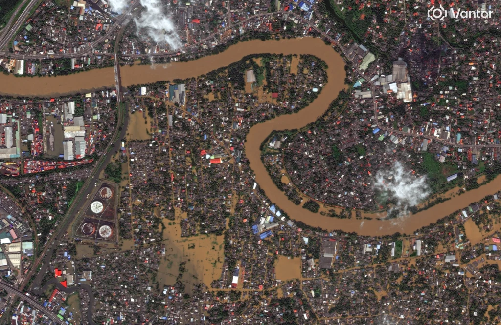 Satelitarne zdjęcie zalanego miasta Colombo na Sri Lance. Na wyspie prezydent ogłosił stan wyjątkowy Zalana rzeka wijąca się przez gęsto zabudowane miasto, widoczne duże obszary podtopione wodą, części infrastruktury miejskiej oraz budynki otoczone brunatną wodą, na niebie miejscami niewielkie chmury.