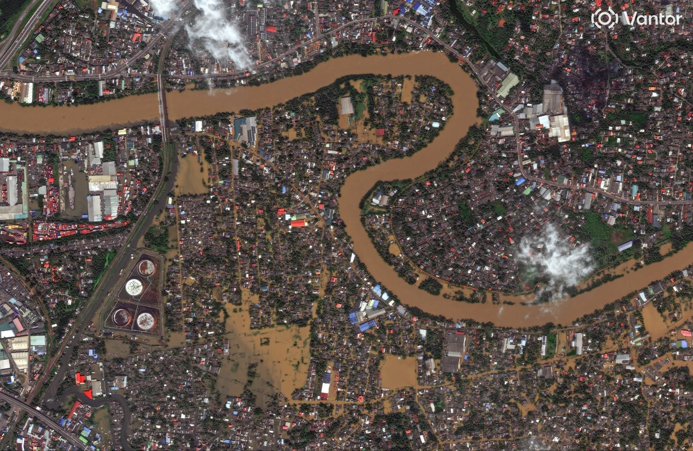 Zalana rzeka wijąca się przez gęsto zabudowane miasto, widoczne duże obszary podtopione wodą, części infrastruktury miejskiej oraz budynki otoczone brunatną wodą, na niebie miejscami niewielkie chmury.