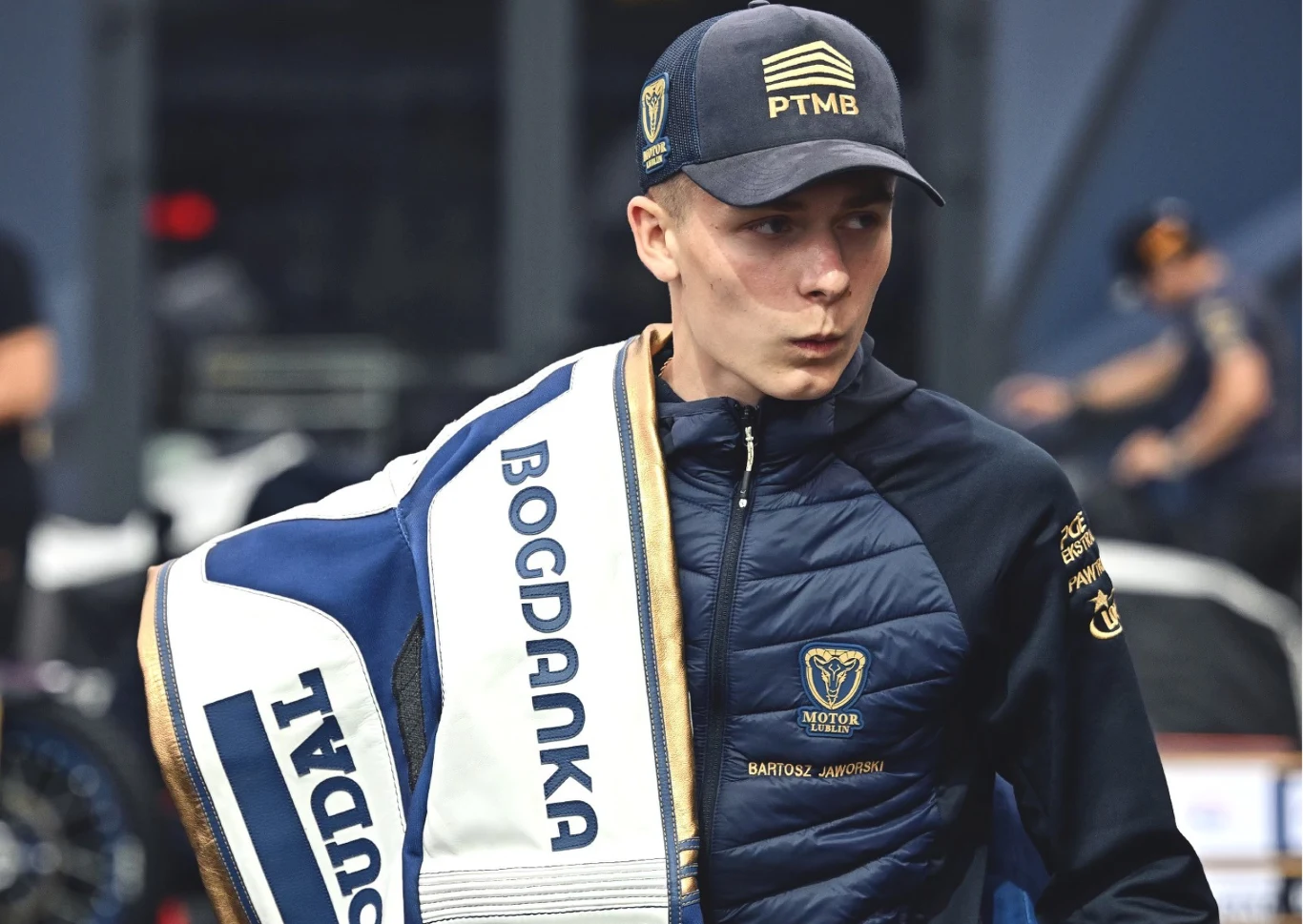Bartosz Jaworski, czyli czas na wychowanka w składzie Motoru Lublin. Młody mężczyzna ubrany w sportową kurtkę i czapkę z daszkiem, trzyma torbę lub specjalny ochraniacz z napisem Bogdanka, wyraźnie skoncentrowany lub zaskoczony, w tle rozmazane postacie i elementy sportowej infrastruktury.