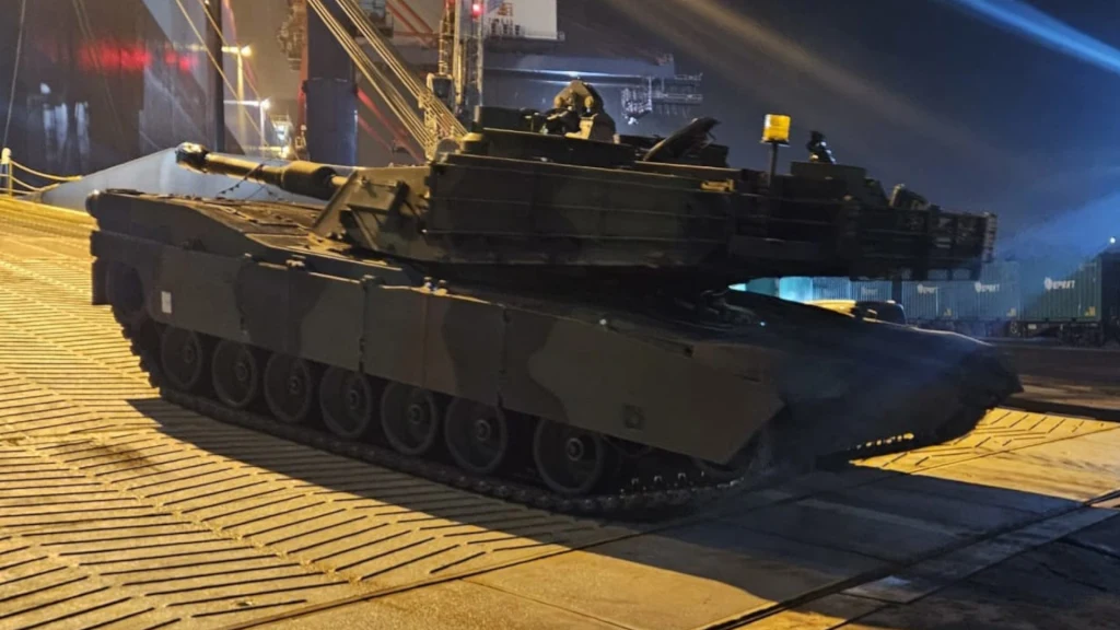 Czołg na rampie wjazdowej oświetlony światłem sztucznym, w tle elementy infrastruktury portowej po zmroku.