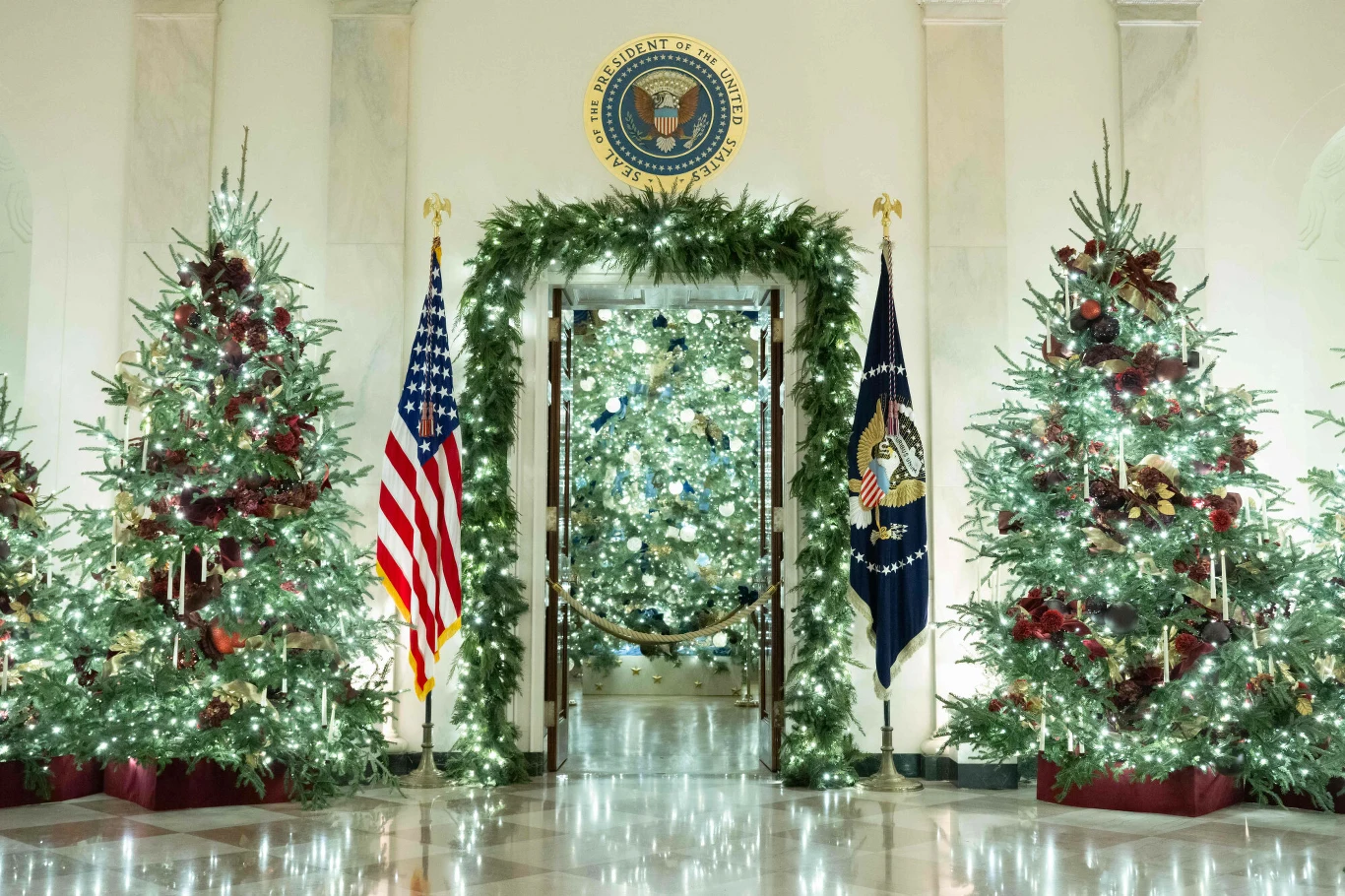 Białe marmurowe wnętrze udekorowane trzema bogato przystrojonymi choinkami, z licznymi lampkami, wstążkami i ozdobami, pośrodku drzwi otoczone girlandą iglastą, amerykańska flaga i flaga prezydenta po bokach, nad drzwiami herb prezydenta USA.