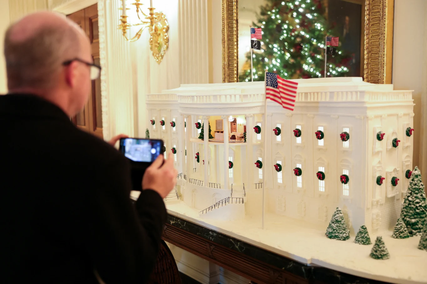 Makieta Białego Domu udekorowana na święta Bożego Narodzenia, ozdobiona miniaturowymi choinkami i wieńcami, z amerykańską flagą na dachu, w tle odbicie choinki świątecznej w lustrze, po lewej mężczyzna fotografujący makietę smartfonem.