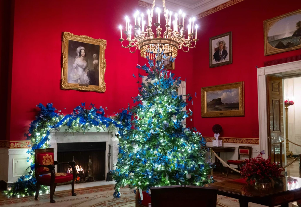 Symbolicznym akcentem tegorocznych dekoracji Białego Domu są niebieskie motyle Pokój z czerwonymi ścianami udekorowany na święta, centralnie umieszczona choinka przystrojona niebieskimi ozdobami i białymi światełkami, w tle kominek z palącym się ogniem oraz żyrandol pod sufitem, na ścianach portrety i obrazy.