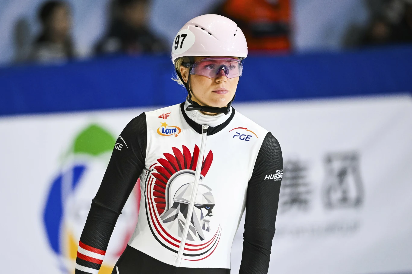 Natalia Maliszewska Zawodniczka w łyżwiarstwie szybkim ubrana w biały strój z czerwonym motywem orła i hełmem, na głowie biały kask z numerem 97, za nią zamazana tablica z napisami i logo.