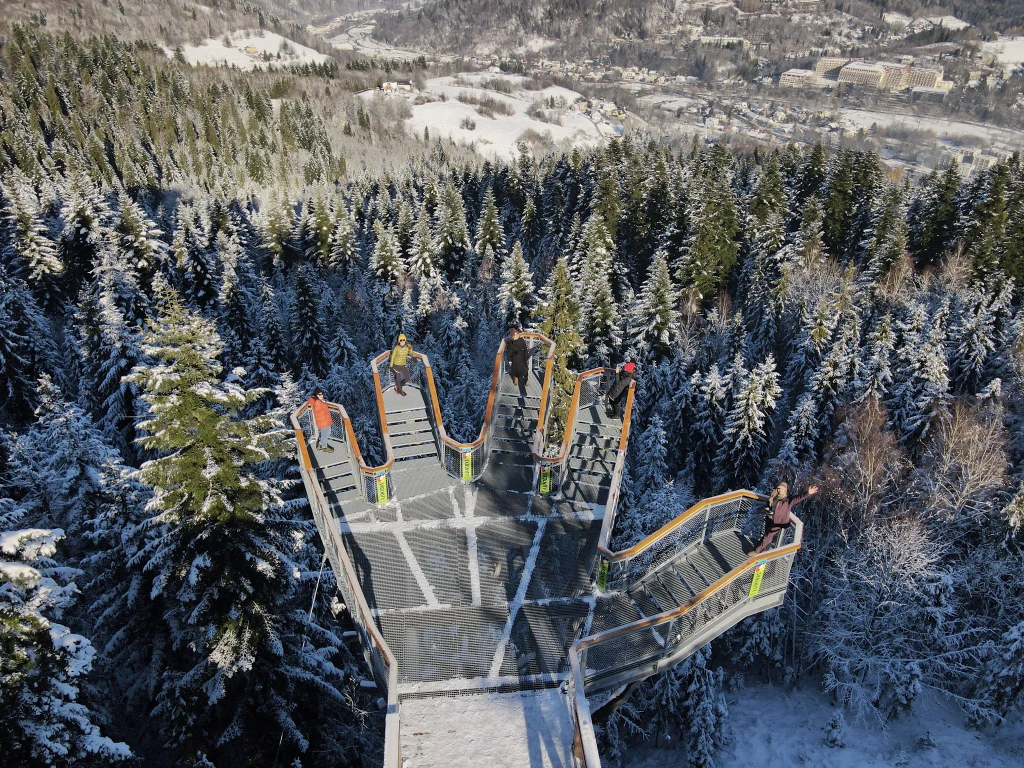 Nowa wieża w Wiśle ma ciekawą konstrukcję - wijące się kładki i balkony widokowe, które układają się w otwartą dłoń. Platforma widokowa w kształcie otwartej dłoni wznosi się ponad zalesionym, zaśnieżonym krajobrazem górskim. Na platformie znajdują się spacerujący ludzie, w tle widok gór, dolin oraz zabudowań miasteczka.