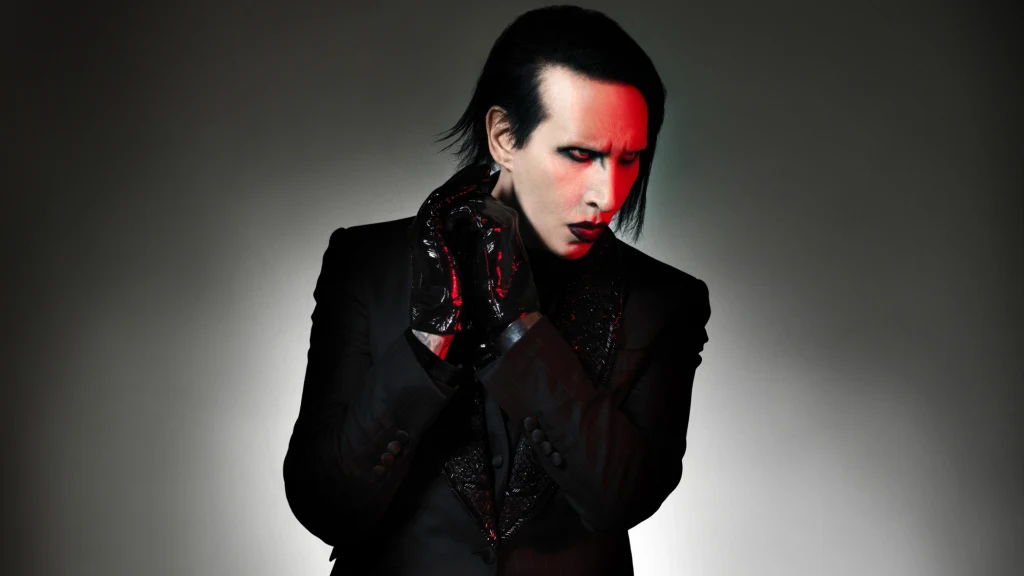 Marilyn Manson Mężczyzna o bladej cerze oraz charakterystycznym, wyrazistym makijażu w ciemnym stroju i czarnych rękawiczkach, zwraca uwagę teatralną pozą oraz ostrym światłem padającym na część twarzy, tworzącym dramatyczny efekt.