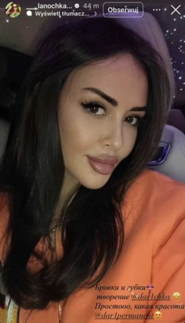 Młoda kobieta z długimi, ciemnymi włosami i starannie zrobionym makijażem, ubrana w pomarańczową bluzę, siedzi w samochodzie na tle tapicerki z efektami świetlnymi.