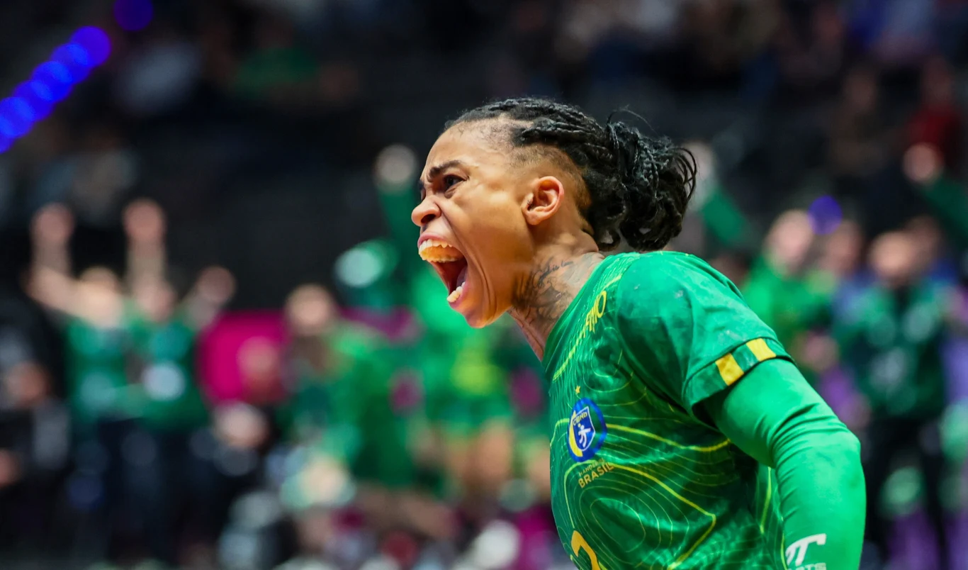 Zawodniczka ubrana w zielony strój sportowy krzyczy z pełną emocji ekspresją na twarzy, w tle zamazana grupa osób, która prawdopodobnie kibicuje podczas wydarzenia sportowego.