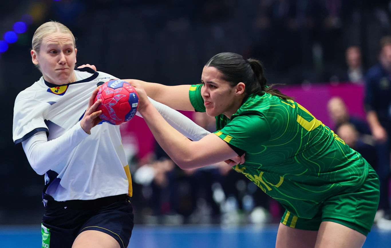 Dwie zawodniczki walczące o piłkę podczas meczu piłki ręcznej, jedna ubrana na biało, druga w zielonym stroju sportowym, dynamiczna akcja i wyraziste emocje na twarzach.