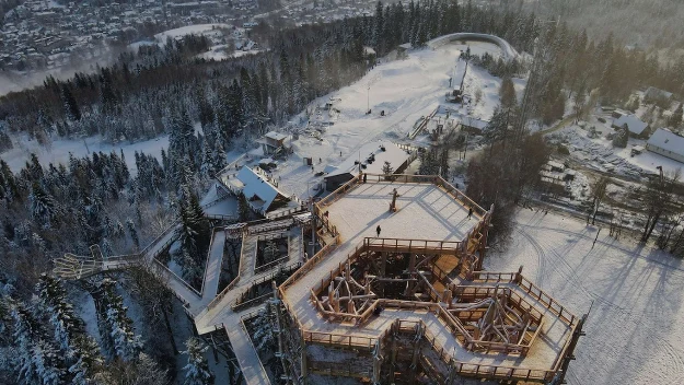 Drewniana wieża widokowa o nowoczesnej konstrukcji otoczona śnieżnym lasem, ścieżki edukacyjne prowadzące przez korony drzew, w oddali panorama miejskiej zabudowy oraz obiekty rekreacyjne pokryte śniegiem.