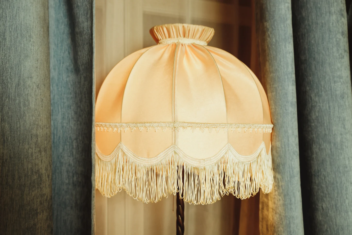 Kremowy abażur lampy w stylu retro z ozdobnymi frędzlami umieszczony przy niebieskich zasłonach, w tle widoczne wnętrze pokoju.