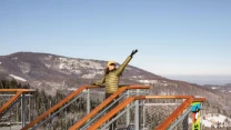 Osoba ubrana w zimową kurtkę i czapkę stoi na platformie widokowej, unosząc jedną rękę ku niebu. W tle rozciągają się ośnieżone góry, lasy oraz bezchmurne niebo. Elementy konstrukcji platformy wykonane są z metalu i drewna, po prawej stronie widoczny j...