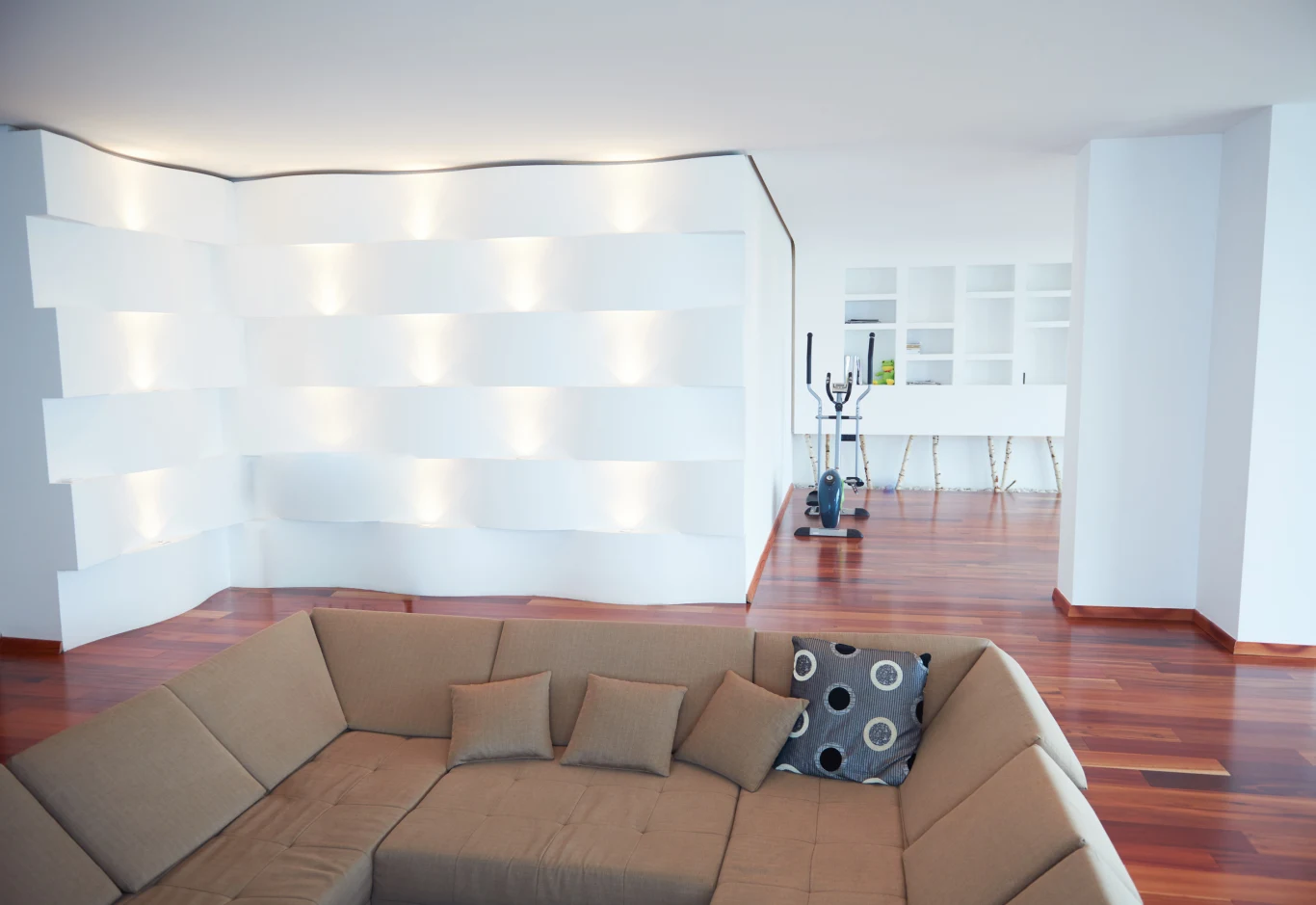 Nowoczesny salon z brązowym narożnikiem oraz poduszkami, jasne ściany i półki z podświetleniem, w tle sprzęt do ćwiczeń przy białym barze kuchennym z wysokimi stołkami, na podłodze drewniane panele.