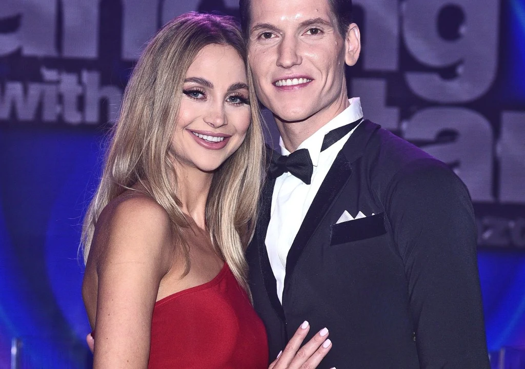 Para elegancko ubranych osób pozuje razem na tle podświetlonej scenografii, kobieta ma na sobie czerwoną suknię bez ramiączek, mężczyzna jest w smokingu z muchą, oboje uśmiechnięci.