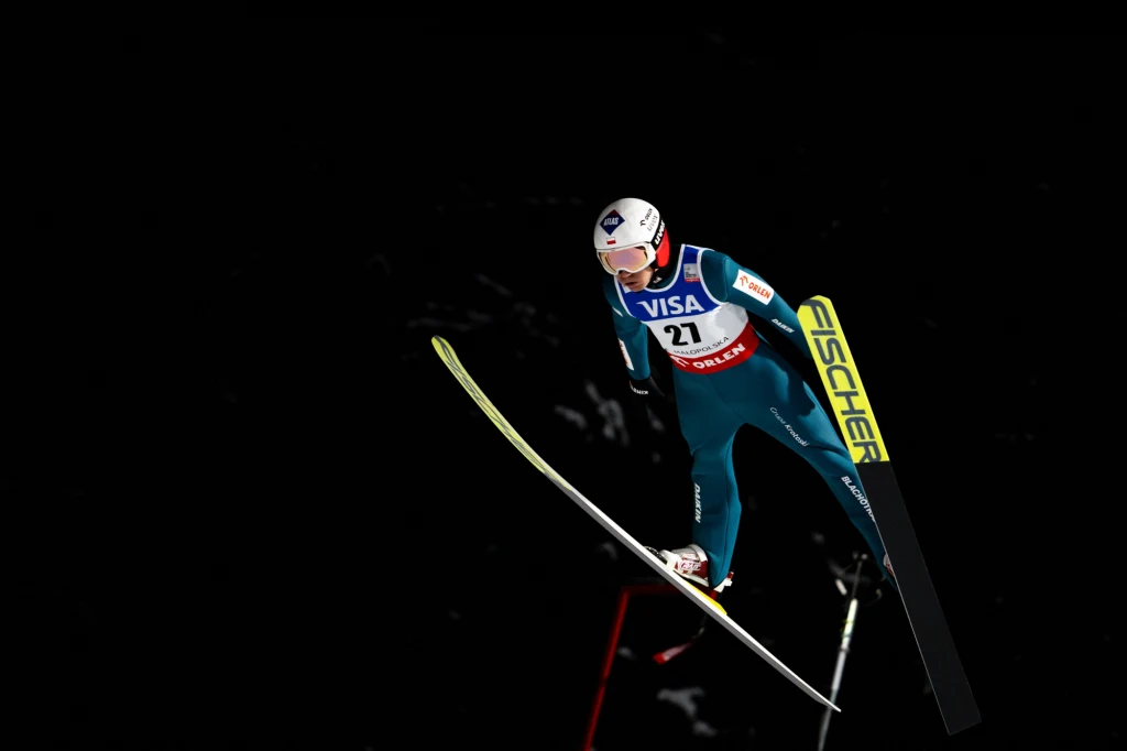 Skoczek narciarski w niebieskim kombinezonie wykonuje skok na tle ciemnego nieba, w pełnym locie z widoczną numeracją startową, kaskiem ochronnym oraz nartami marki Fischer.