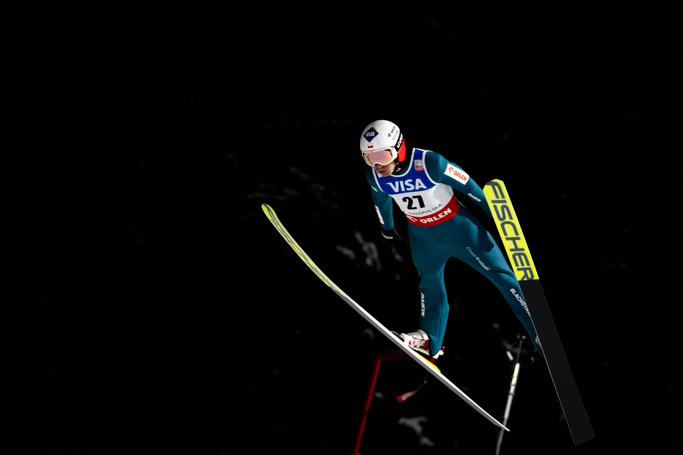 Skoczek narciarski w niebieskim kombinezonie wykonuje skok na tle ciemnego nieba, w pełnym locie z widoczną numeracją startową, kaskiem ochronnym oraz nartami marki Fischer.