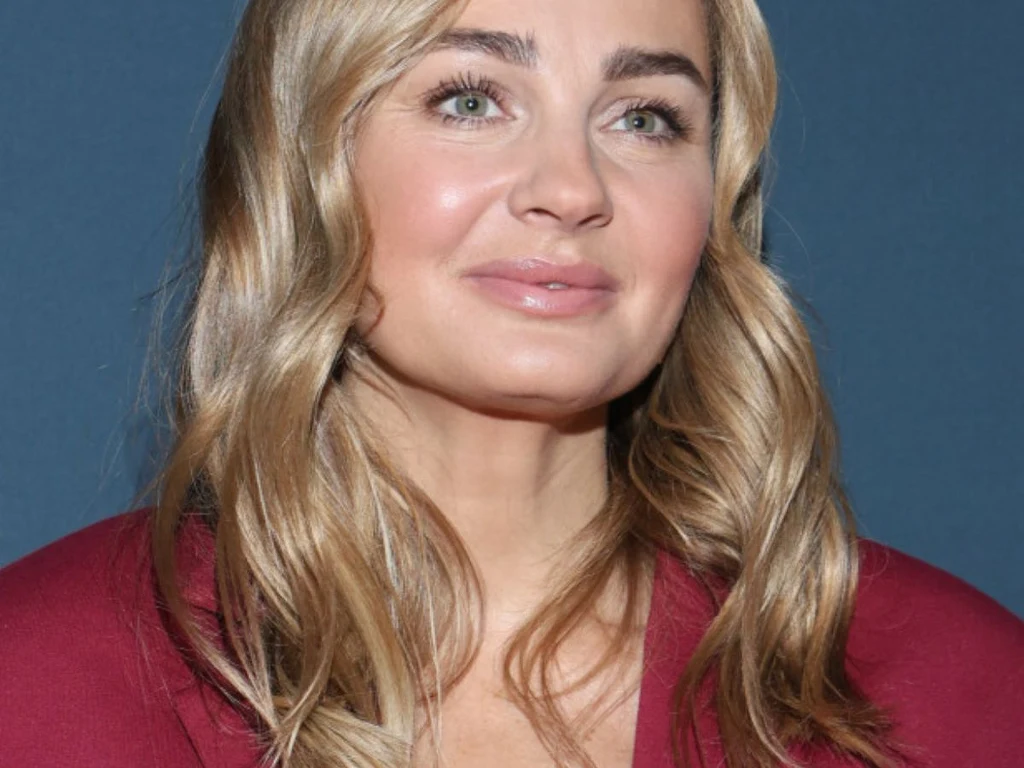 Uśmiechnięta kobieta o blond włosach, ubrana w bordowy strój, na tle jednolitej niebieskiej ściany.