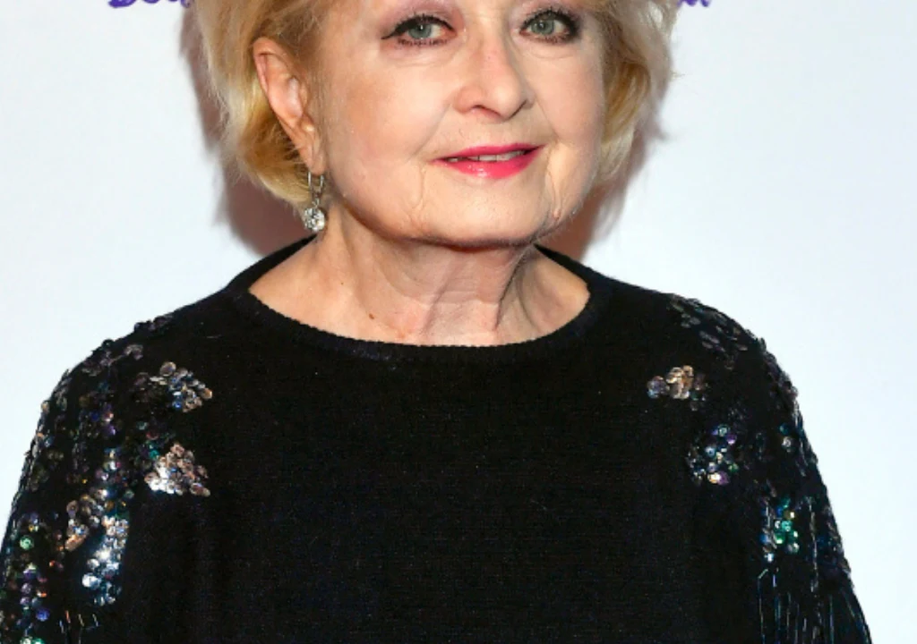 Starsza kobieta o jasnych włosach, ubrana w czarną, ozdobioną cekinami bluzkę, stoi na tle jasnego tła. Uśmiecha się lekko i nosi subtelne kolczyki.