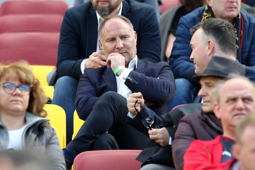 Dariusz Adamczuk podczas meczu Pogoni Szczecin Grupa osób siedząca na stadionowych trybunach, mężczyzna w garniturze w centrum ujęcia trzyma dłoń przy ustach i rozmawia z innym mężczyzną, wokół niego inne osoby skupione na rozmowie lub wydarzeniu.